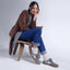 Woman sitting on a chair in front of a white wall, wearing a brown jacket, checkered scarf, blue jeans, and white socks with the Balboa Sneaker in slate nubuck