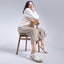Woman sitting on a wood chair against a white wall, wearing the Balboa Sneaker in white leather paired with beige pants and a white sweater