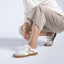 Person squatting down against a white wall, wearing the Balboa Sneaker in white leather paired with beige pants and a white sweater