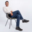 Man sitting on a chair wearing a white sweater, blue jeans, and black shoes against a white background