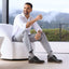 Man sitting on a white chair wearing a white shirt, gray pants, and featherlight founder black shoes with a scenic background.