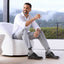 Man sitting on a white chair wearing a white shirt, gray pants, and featherlight founder black shoes with a scenic background.