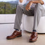 Person wearing featherlight founder brown leather shoes and gray pants sitting on a white chair with a scenic background.