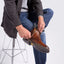 A man sitting on a metal stool puts on the featherlight lace-up boot while wearing blue jeans and a dark jacket against a light gray background.