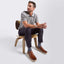A man sits on a wooden chair wearing a gray short-sleeve polo shirt, dark trousers, gray socks, and Featherlight Loafers against a light gray studio background.