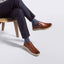 A person seated on a wooden stool wearing Featherlight loafers with beige soles, dark navy trousers, and gray socks against a clean light gray studio background.