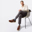 A man sits on a modern chair against a white studio background, wearing Featherlight Napa in Bourbon Leather with light beige soles, dark gray trousers, and a cream long-sleeve shirt.