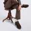 A person seated on a wooden stool wearing brown trousers, a dark knit sweater, white socks, and the Featherlight Founder against a light gray studio background.