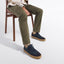 A person seated on a wooden chair wearing olive green pants and Hayes Sneaker in dark navy nubuck with gum soles