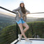 Woman standing on a balcony with a scenic view, wearing a white cap, patterned jacket, denim shorts, and featherlight Laguna textured pearl sneakers.