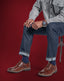 Person wearing brown leather shoes, blue jeans, and a gray shirt against a red background