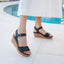 Portola Two-Strap Wedge Black Leather worn by a person standing on a tiled floor near a pool.
