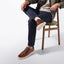 A person seated on a wooden chair wearing dark blue jeans, and Presidio Sneaker with off-white soles against a clean white studio background.
