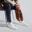 A person seated on a chair wearing Presidio Sneaker in white leather and holding the Antique Brown Leather version against a light gray studio background.