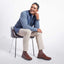 Man sitting on a chair wearing a blue sweater, beige pants, and brown shoes against a white background