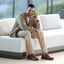 A man is sitting on a white couch, talking on a phone in a modern living room, wearing Rafael Slip-On cognac leather shoes.