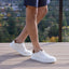 Person wearing Sunset Sneaker 2.0 White Leather sneakers with brown accents on a wooden deck.